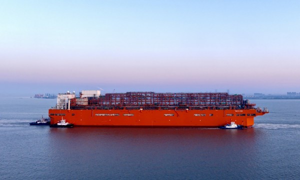 Nave cargo arancione scortata da due rimorchiatori in porto al tramonto.