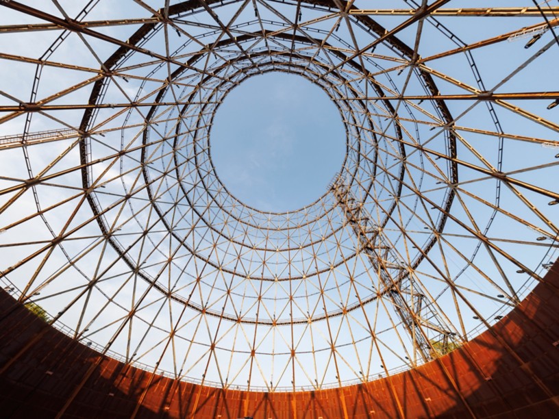 Circular metal structure viewed from below, with a network of steel beams forming a geometric lattice against the blue sky. Warm sunset light highlights the lower parts of the structure.