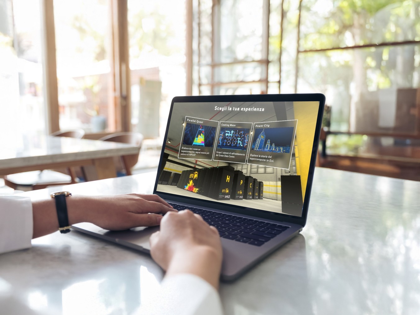 Persona seduta a un tavolo usa un laptop con un’interfaccia digitale che mostra diverse opzioni di esperienza; ambiente luminoso con grandi finestre sullo sfondo.