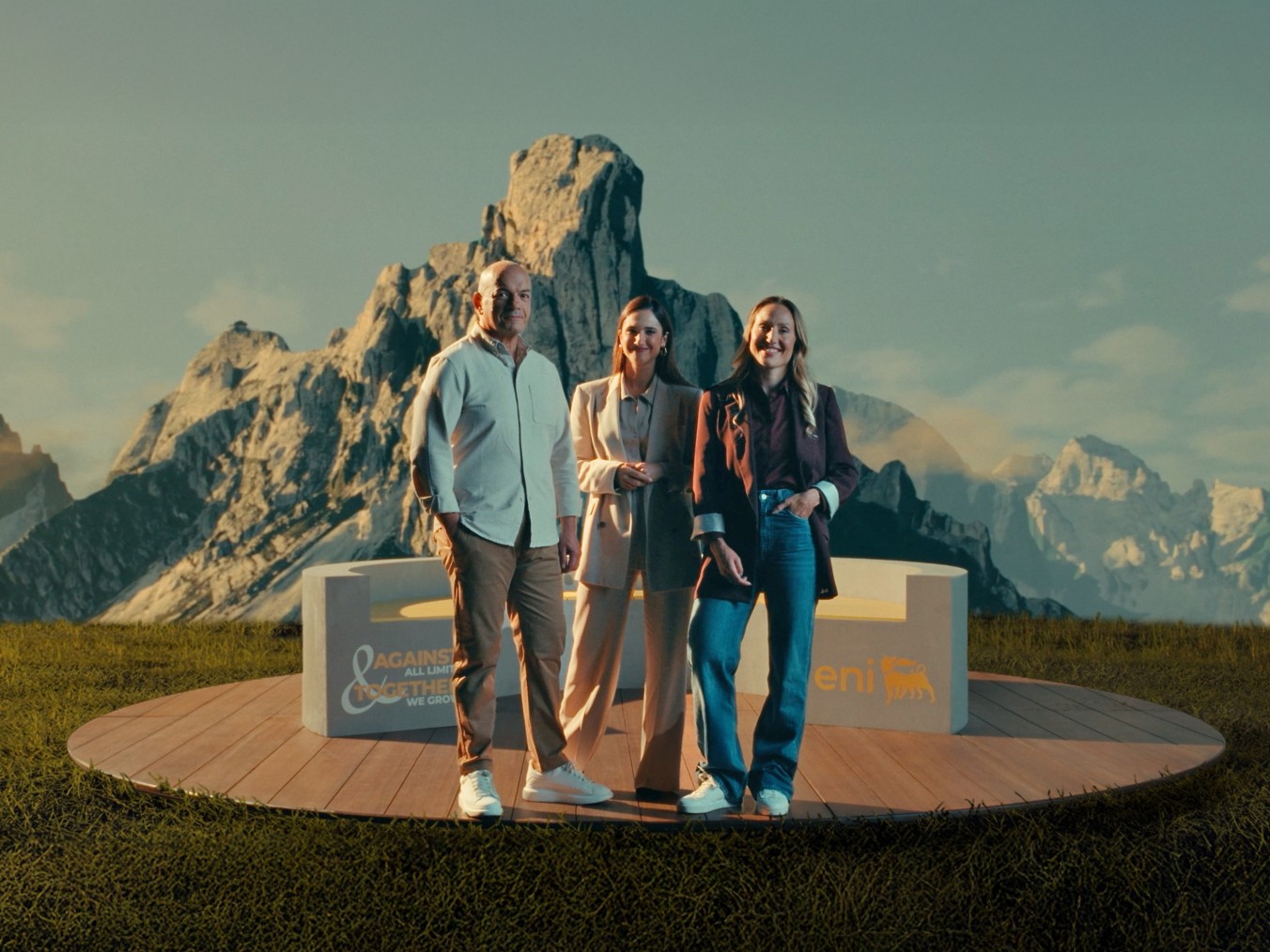 Tre persone in piedi su una piattaforma circolare di legno, davanti a un paesaggio montano suggestivo. Sorridono e indossano abiti eleganti ma informali; sulla piattaforma si leggono i loghi Eni e “Against all limits, together we grow”.