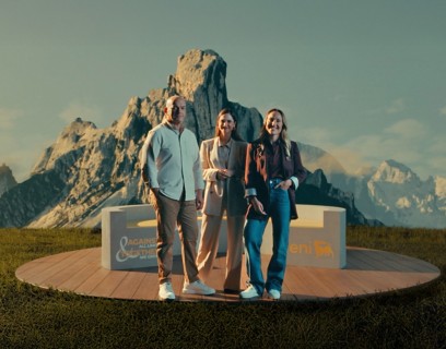 Tre persone in piedi su una piattaforma circolare di legno, davanti a un paesaggio montano suggestivo. Sorridono e indossano abiti eleganti ma informali; sulla piattaforma si leggono i loghi Eni e “Against all limits, together we grow”.