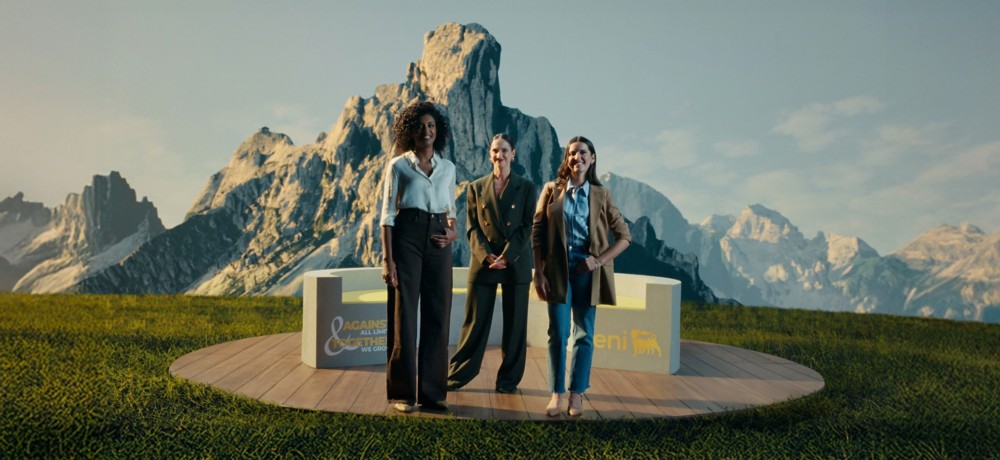 Tre donne in piedi su una piattaforma circolare di legno, immerse in un paesaggio alpino con montagne maestose sullo sfondo. Indossano abiti eleganti e professionali, accanto a una struttura con i loghi “Eni” e “Against All Limits – Together We Grow”.