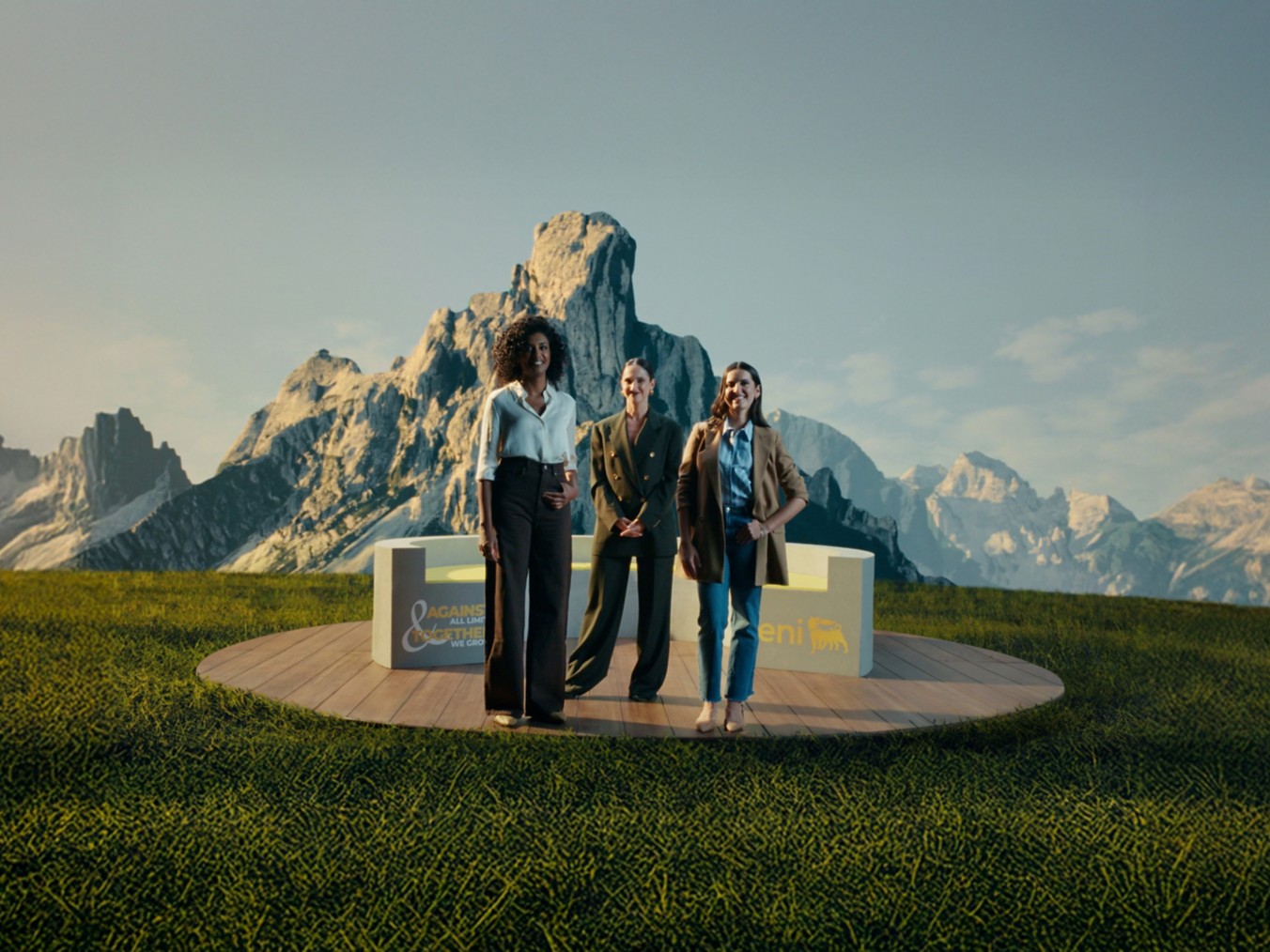 Tre donne in piedi su una piattaforma circolare di legno, immerse in un paesaggio alpino con montagne maestose sullo sfondo. Indossano abiti eleganti e professionali, accanto a una struttura con i loghi “Eni” e “Against All Limits – Together We Grow”.