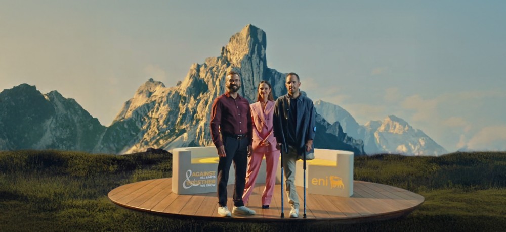 Tre persone su una pedana di legno davanti a montagne illuminate; due uomini e una donna posano sorridendo accanto a sedute con i loghi “eni” e “AGAINST ALL LIMITS TOGETHER WE GROW”.