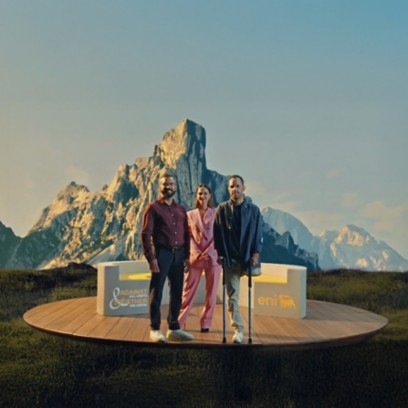 Tre persone su una pedana di legno davanti a montagne illuminate; due uomini e una donna posano sorridendo accanto a sedute con i loghi “eni” e “AGAINST ALL LIMITS TOGETHER WE GROW”.