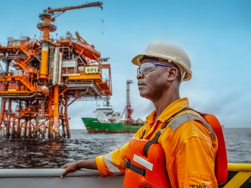 Operaio di profilo su una nave fissa davanti a sé, sullo sfondo l'impianto offshore del campo di Nené