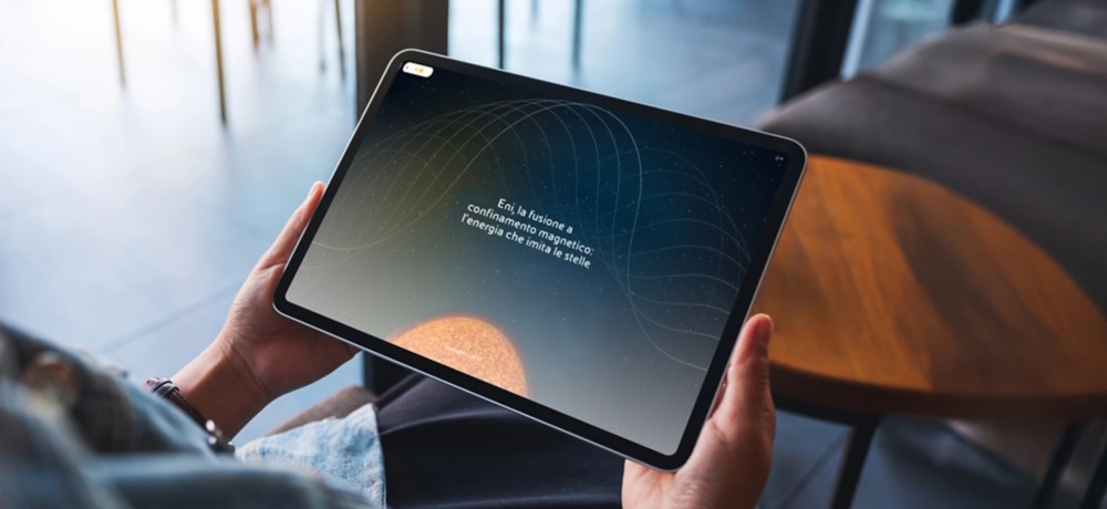 Persona con tablet  seduta in un bar, sullo schermo un'interfaccia del longform sulla fusione Eni