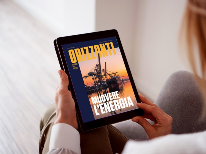 Tablet bianco inclinato su sfondo azzurro mostra la copertina della rivista “Orizzonti” con una nave cargo in porto al tramonto e il titolo “Muovere l’energia”.