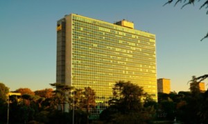 Vista esterna del Palazzo Mattei di Eni a Roma, un edificio alto con una facciata composta da numerose vetrate riflettenti, illuminate dalla luce calda del tramonto. Davanti al palazzo si vedono alberi e un’area verde, con una strada e un autobus in basso, sotto un cielo limpido.