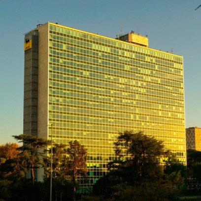 Vista esterna del Palazzo Mattei di Eni a Roma, un edificio alto con una facciata composta da numerose vetrate riflettenti, illuminate dalla luce calda del tramonto. Davanti al palazzo si vedono alberi e un’area verde, con una strada e un autobus in basso, sotto un cielo limpido.