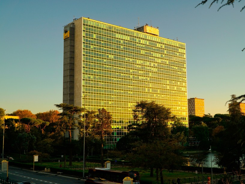 Vista esterna del Palazzo Mattei di Eni a Roma, un edificio alto con una facciata composta da numerose vetrate riflettenti, illuminate dalla luce calda del tramonto. Davanti al palazzo si vedono alberi e un’area verde, con una strada e un autobus in basso, sotto un cielo limpido.