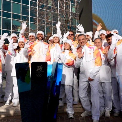 Gruppo di persone in uniforme bianca dei Giochi Milano Cortina 2026 che sorridono e salutano davanti a un edificio moderno, con la torcia o il simbolo dell’evento in primo piano.
