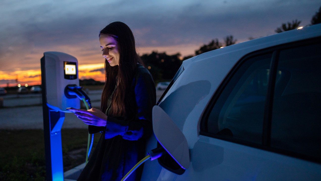 Ragazza guarda il cellulare al tramonto mentre ricarica la macchina elettrica