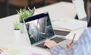 Persona seduta a una scrivania che lavora al computer portatile; sullo schermo è visibile una schermata con la scritta "Eni for 2024". Sulla scrivania ci sono piante, post-it colorati e un quaderno.
