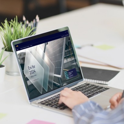 Persona seduta a una scrivania che lavora al computer portatile; sullo schermo è visibile una schermata con la scritta "Eni for 2024". Sulla scrivania ci sono piante, post-it colorati e un quaderno.