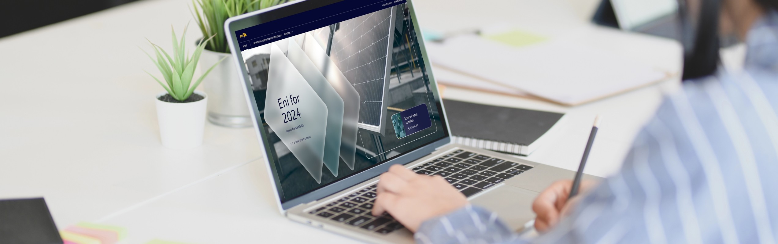 Persona seduta a una scrivania che lavora al computer portatile; sullo schermo è visibile una schermata con la scritta "Eni for 2024". Sulla scrivania ci sono piante, post-it colorati e un quaderno.