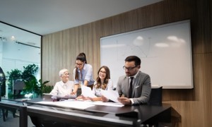 Gruppo di colleghi in riunione in ufficio: una donna in piedi mostra un documento agli altri seduti al tavolo, mentre discutono e sorridono
