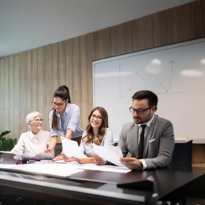 Group of colleagues in a meeting room: a woman standing shows a document to others seated at the table, while they discuss and smile