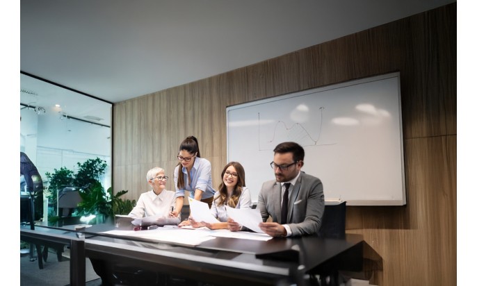 Group of colleagues in a meeting room: a woman standing shows a document to others seated at the table, while they discuss and smile