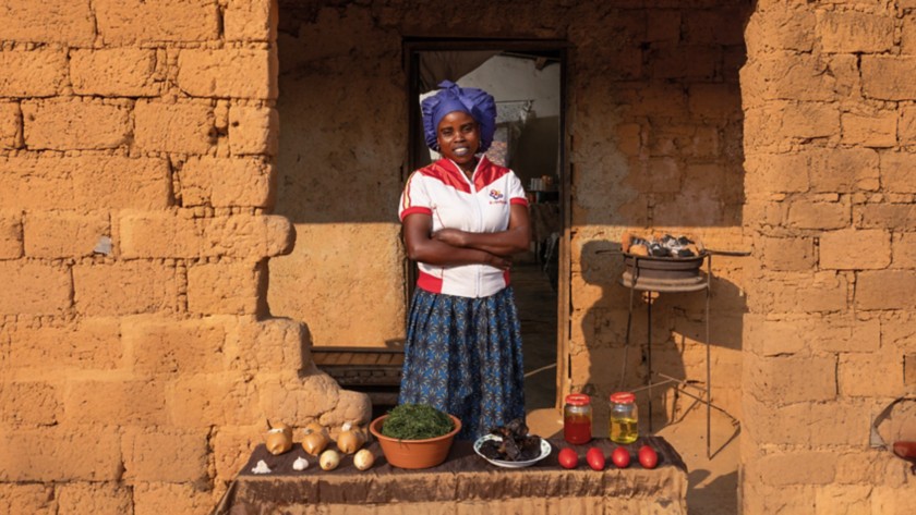 Woman standing in front of the entrance of an earthen house with her arms crossed. In front of her is a table displaying various local foods, including onions, herbs, tomatoes, jars of oil, and other ingredients. To her right there is a small charcoal brazier.