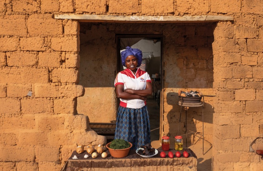 Woman standing in front of the entrance of an earthen house with her arms crossed. In front of her is a table displaying various local foods, including onions, herbs, tomatoes, jars of oil, and other ingredients. To her right there is a small charcoal brazier.