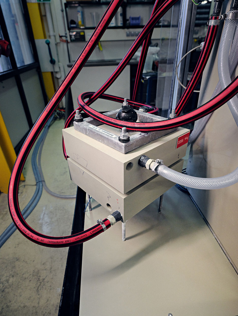 Ein grauer Kasten. Drei rot-schwarze Schläuche führen hinein, außerdem ein transparenter Wasserschlauch.