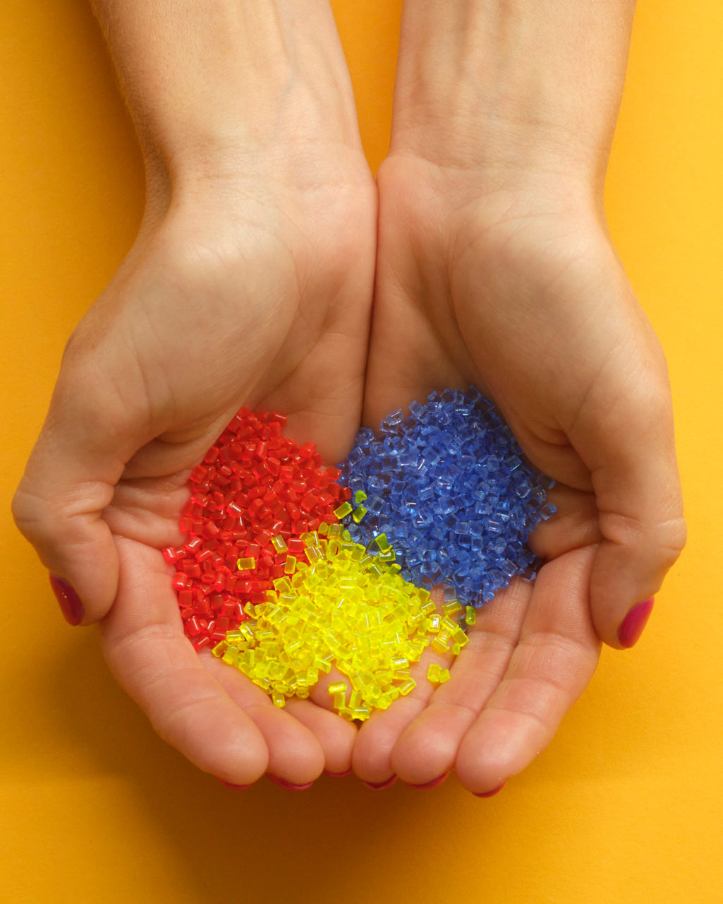 Two female hands hold color-separated PA12 pellets in blue, yellow, and red.