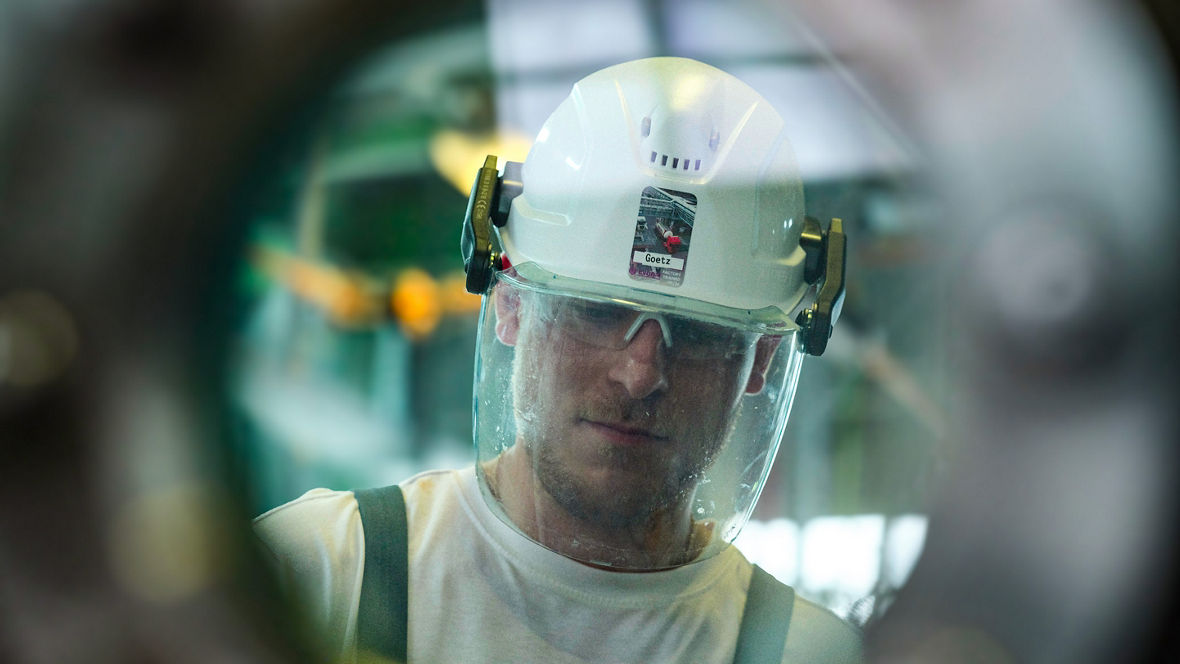 An Evonik employee wearing a helmet and safety glasses looks through a round viewing window.