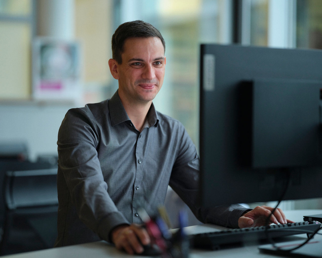 Thomas Asche is sitting at a computer workstation and smiling.