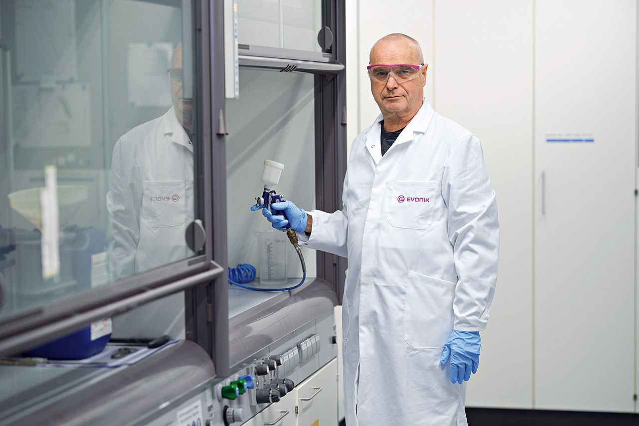 Chemotechniker Eckhard Just im Kittel mit Spritzpistole, Handschuhen und Schutzbrille an einem Versuchsschrank.