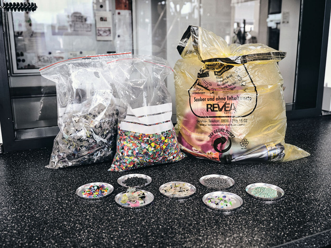 Three bags of plastic waste. In front of them are seven trays with separated plastic waste.