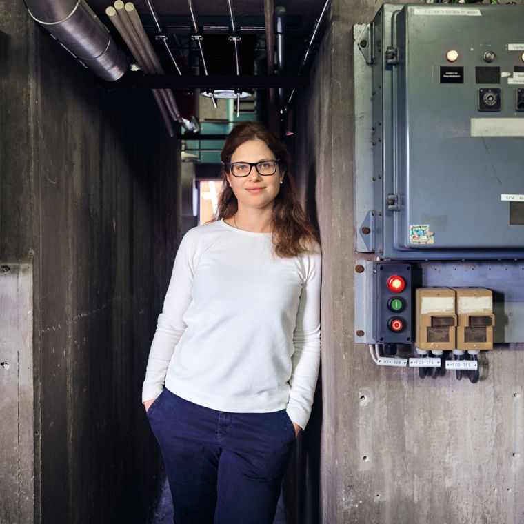 Evonik-Forscherin Jutta Malter lehnt an einer Wand, darüber eine große Uhr, neben ihr Schaltkästen.