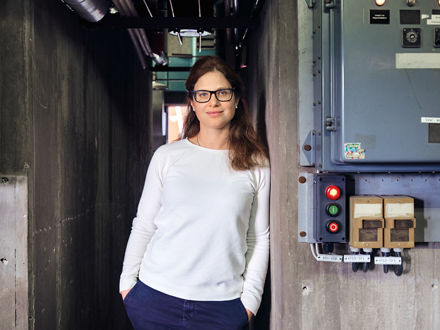 Evonik-Forscherin Jutta Malter lehnt an einer Wand, darüber eine große Uhr, neben ihr Schaltkästen.