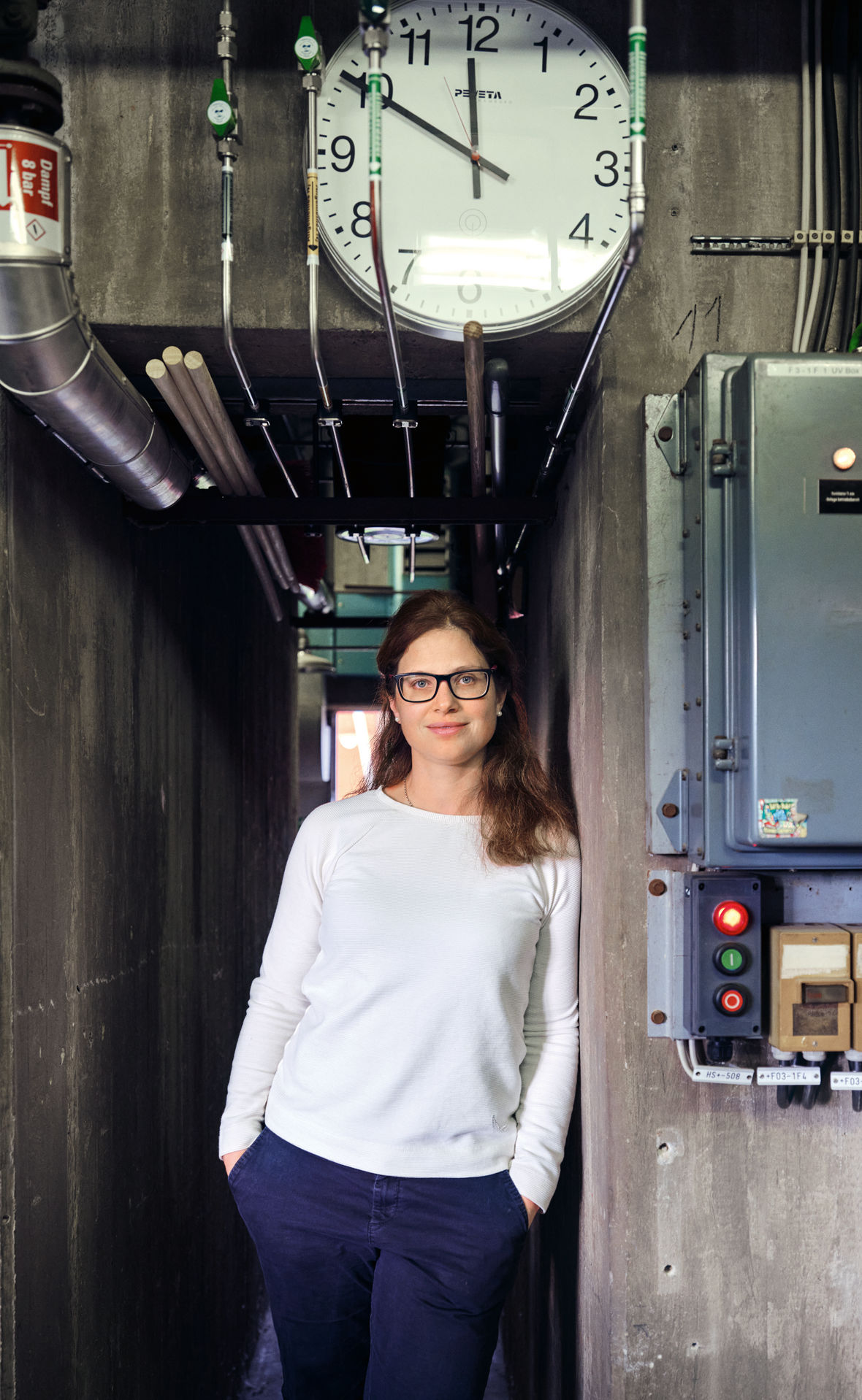 Evonik-Forscherin Jutta Malter lehnt an einer Wand, darüber eine große Uhr, neben ihr Schaltkästen.