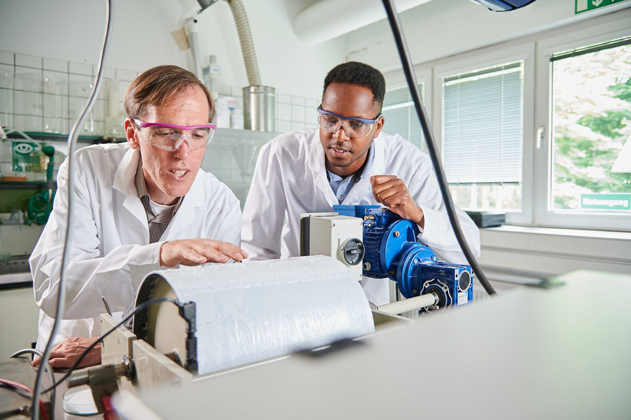 Two men in lab coats stand behind a roughly 50-centimeter-wide (20-inch) roller.
