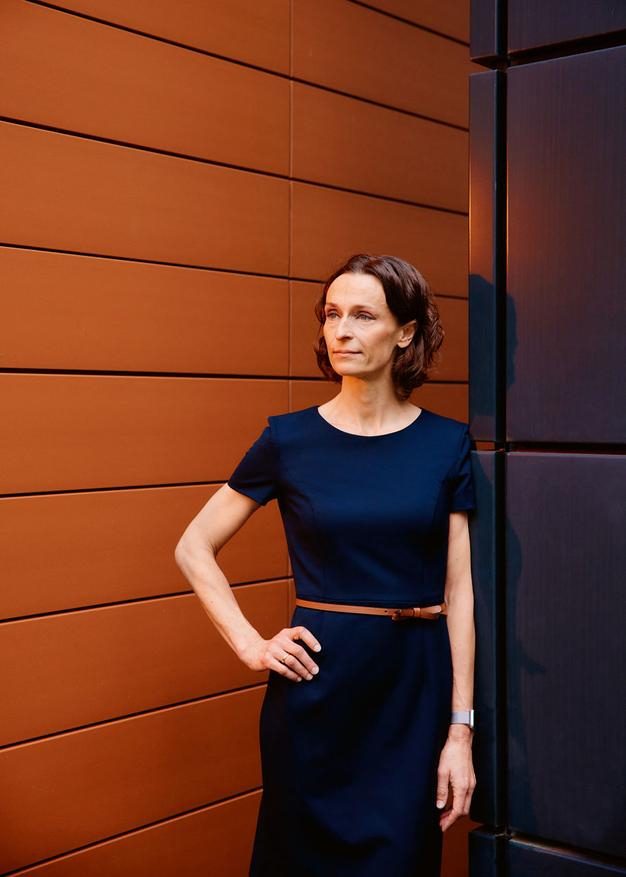 Heike tom Dieck is leaning against a blue corner in front of a wood-coloured wall. She is wearing a blue dress and looks past the camera on the left.