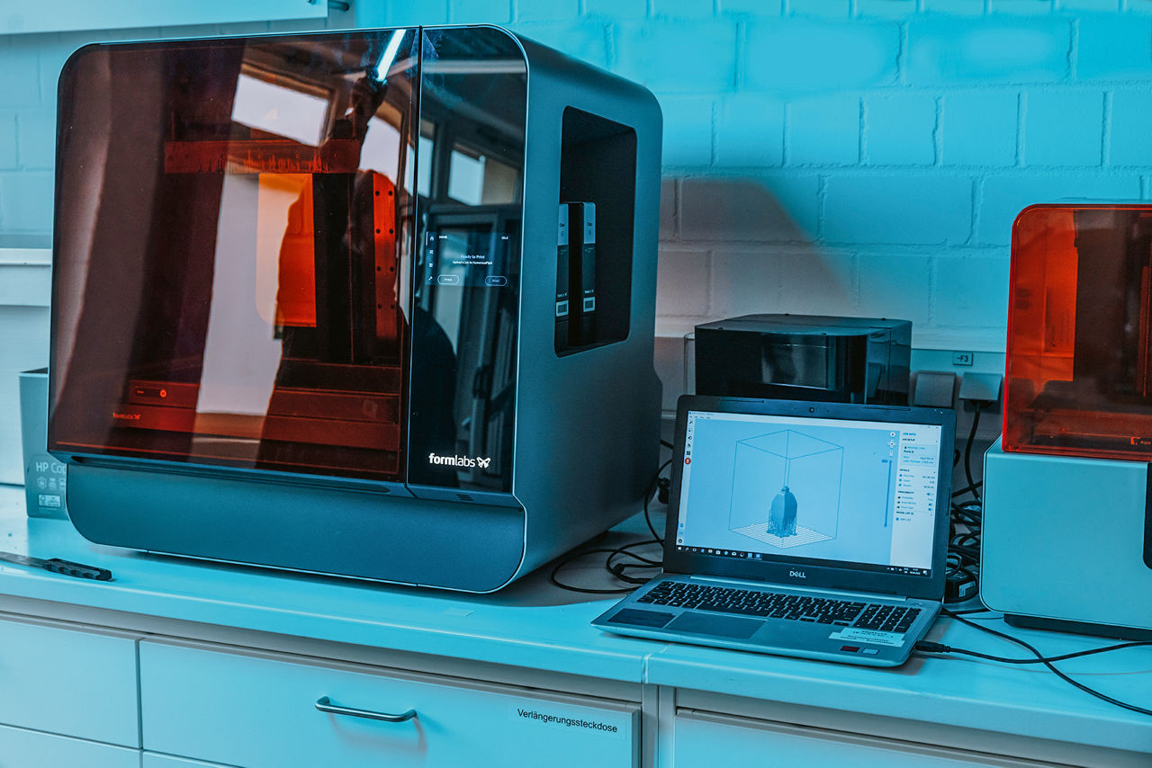 Ein 3D-Drucker steht neben einem Laptop. Der Drucker ist etwa 80 Zentimeter hoch und hat eine glänzend rote Front.