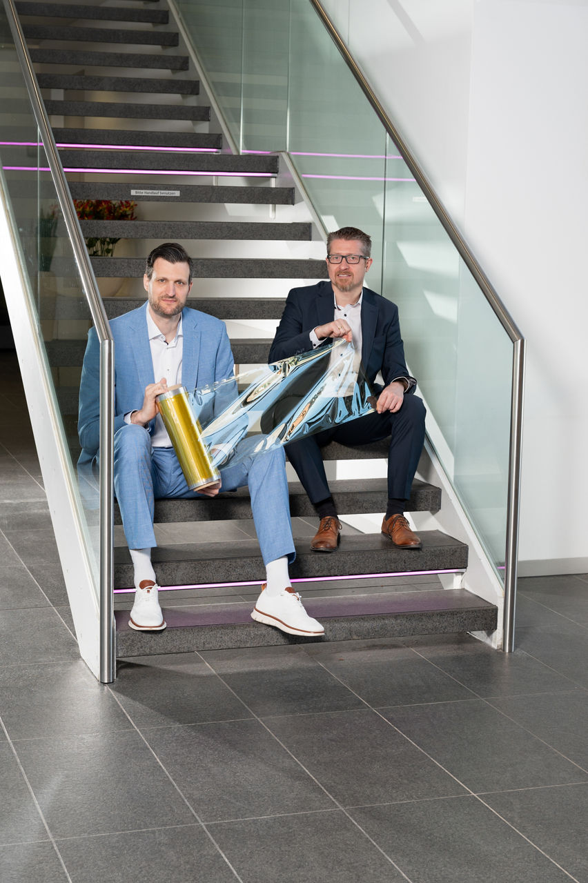 Christian Schnitzer und Christian Däschlein sitzen gemeinsam auf einer modernen Treppe. Sie halten eine Rolle der Membran in den Händen.