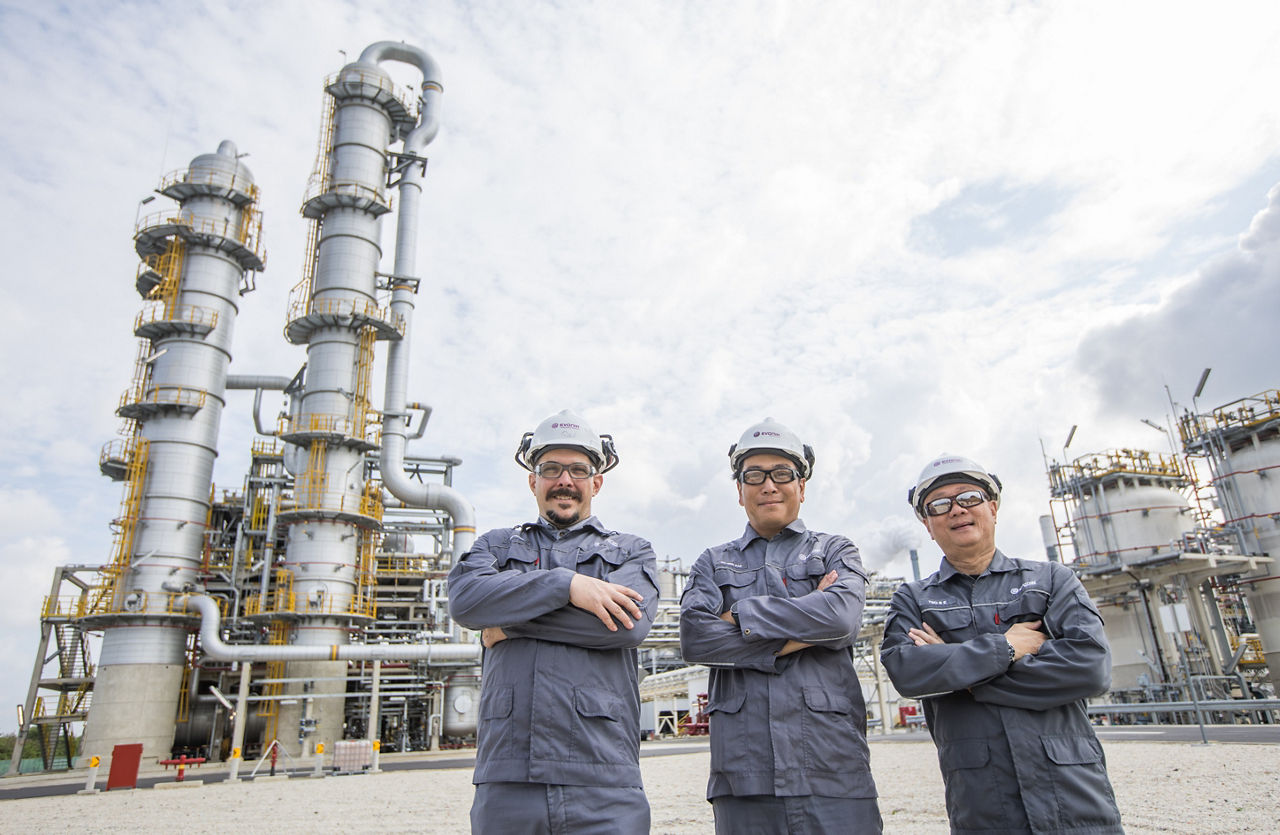 Drei Männer in Arbeitsausstattung mit verschränkten Armen vor den Kolonnen der Anlage.