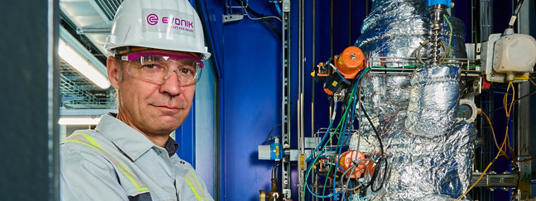 Mann mit Schutzhelm und Arbeitskleidung steht vor isoliertem, komplexem Industrieanlagenrohr.