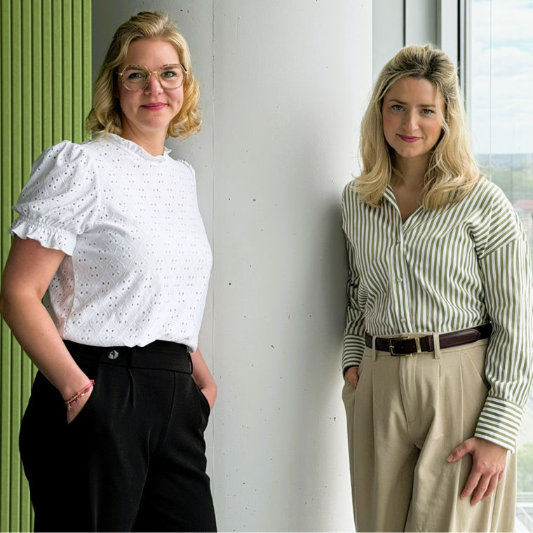 Two women standing by a large window with a city view, one wearing a white blouse and the other striped.
