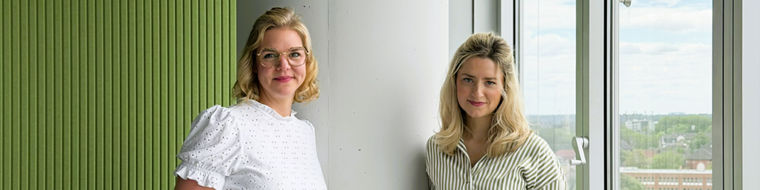 Two women standing by a large window with a city view, one wearing a white blouse and the other striped.