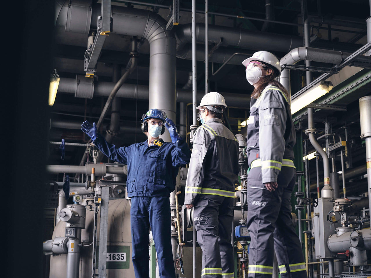 Drei menschen mit Helmen, Arbeitskleidung und Mundschutz in der Anlage.