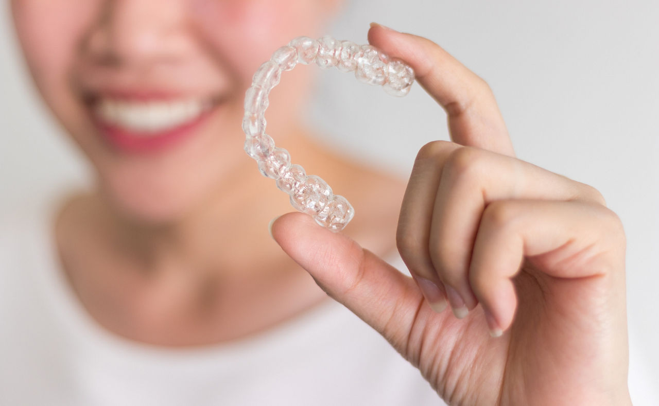 A smiling person holding a clear dental aligner between their fingers against a white background.