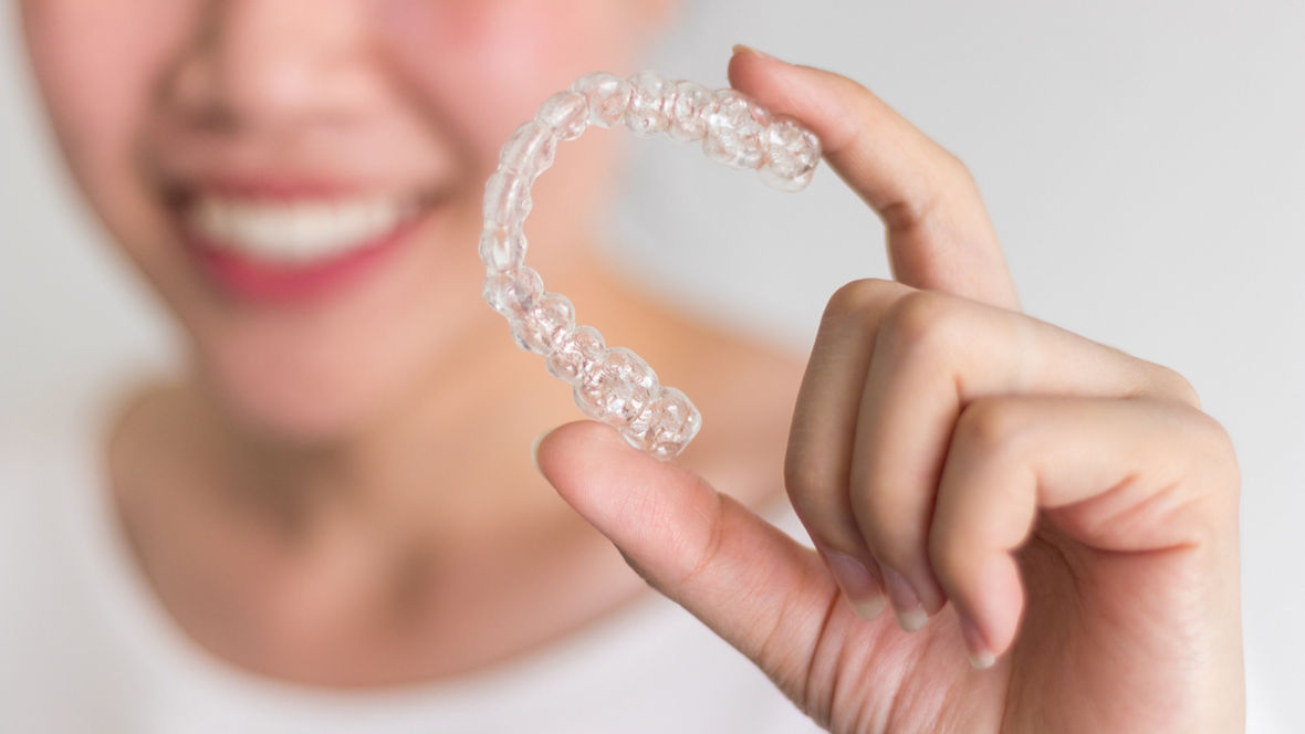 A smiling person holding a clear dental aligner between their fingers against a white background.