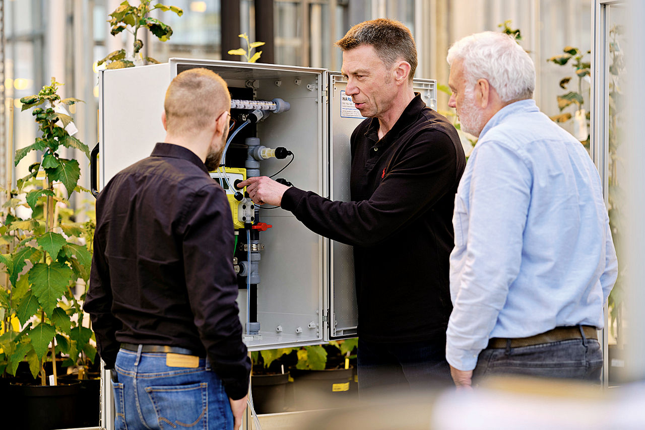 Drei Männer stehen vor einem grauen Kasten, in dem die tTechnik installiert ist.