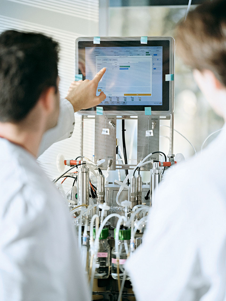 Zwei Labormitarbeiter schauen auf einen Bildschirm, der eine zeigt darauf. Zu sehen ist darunter auch ein kleiner Fermenter.