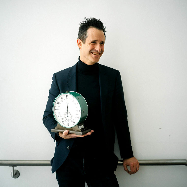 Thorsten Schumm hält eine große analoge Stoppuhr auf der rechten Hand und lacht.