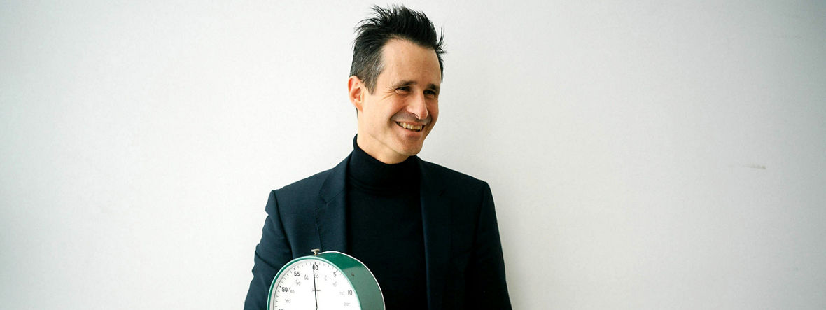 Thorsten Schumm holds a large analog stopwatch in his right hand and laughs. Thorsten Schumm holds a test tube containing a tiny amount of green liquid in his hand.