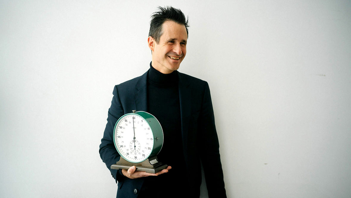 Thorsten Schumm hält eine große analoge Stoppuhr auf der rechten Hand und lacht.